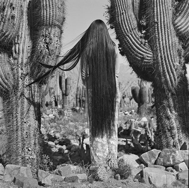 Mujer de espaldas con el cabello larguísimo entre dos cactus, en blanco y negro